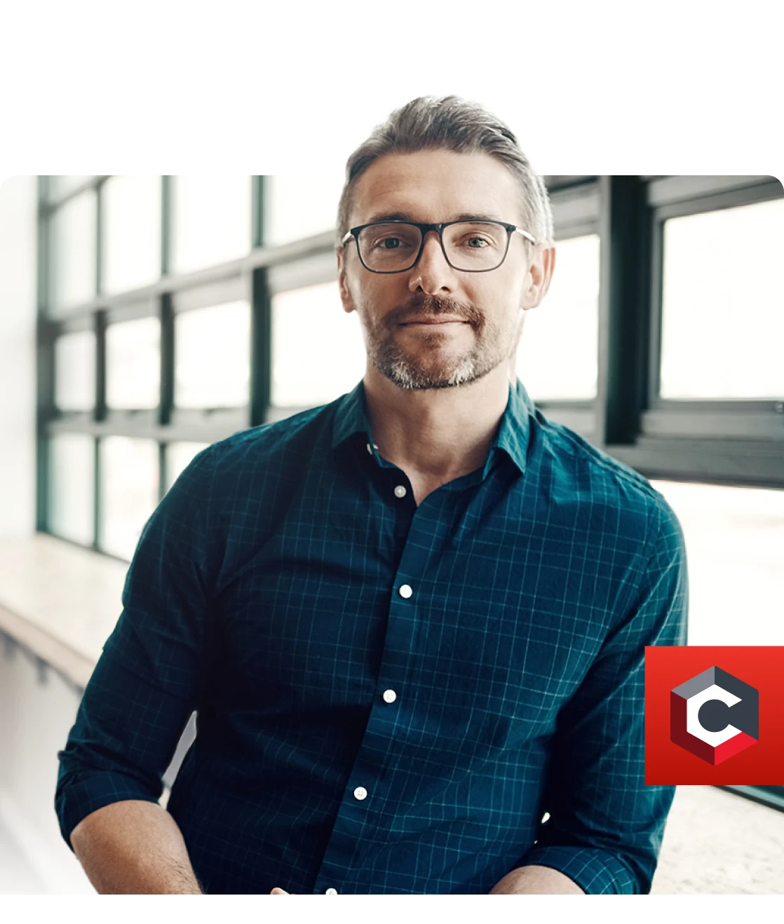 Portrait of a man with glasses wearing a teal checkered shirt in a bright office setting.